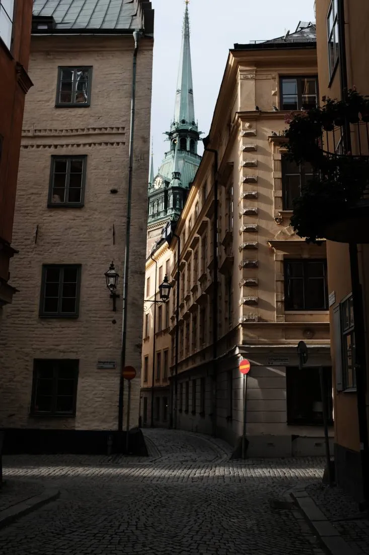 A narrow cobbled lane in Gamla stan with the green copper spire of the German Church between sunlit facades