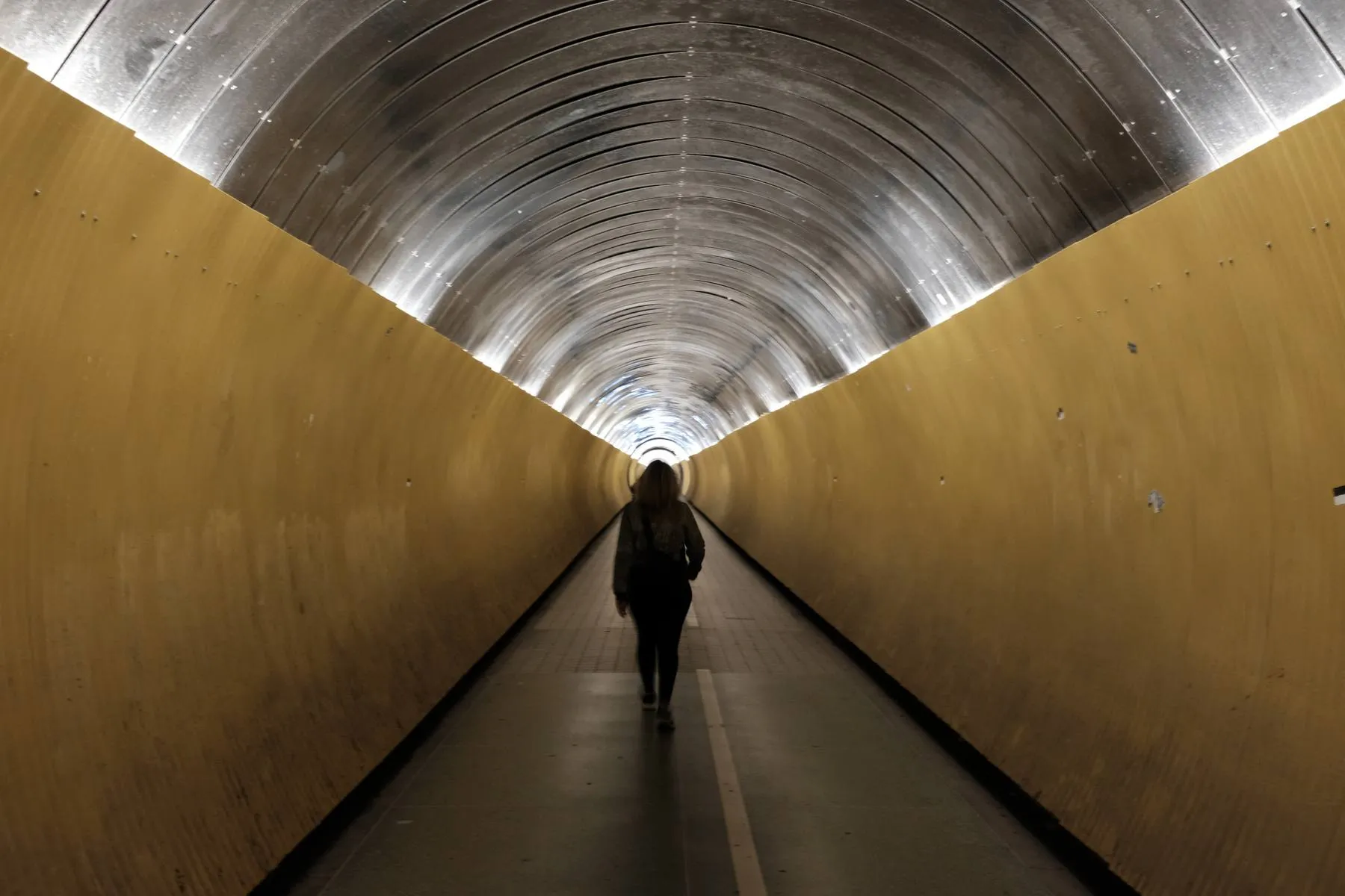 A person walking away through a long yellow tunnel with a polished steel arch, towards daylight